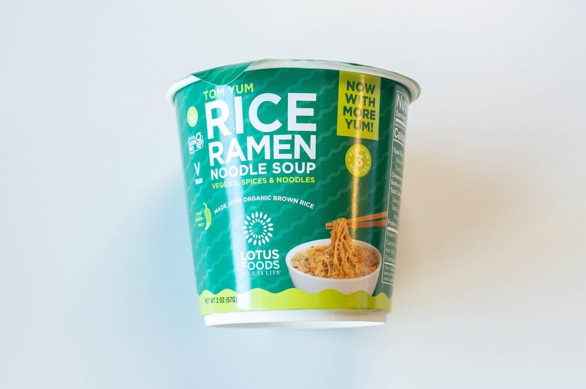 The green and white cup of Lotus Foods Tom Yum Rice Ramen Noodle Soup on a white surface, showing TOM YUM in small orange lettering at the top, RICE RAMEN NOODLE SOUP in bold white block lettering, VEGGIES, SPICES & NOODLES in yellow, "Made with Organic Brown Rice" tag, a small illustration of a white bowl of noodles with chopsticks, and a green NOW WITH MORE YUM! starburst in the upper right.
