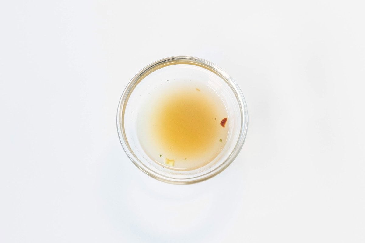 A small clear glass ramekin of the Lotus Foods Tom Yum Rice Ramen Noodle Soup broth on a white surface, showing a very pale yellow-amber liquid with a small red chili flake visible floating in it.