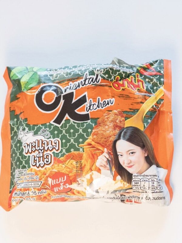 The orange and green package of MAMA Oriental Kitchen Instant Noodles Beef Panang Curry on a white surface, showing the OK Oriental Kitchen logo at the top left, the MAMA brand mark at the top right, and a model holding chopsticks of finished orange-red noodles next to the Thai product name in red script.