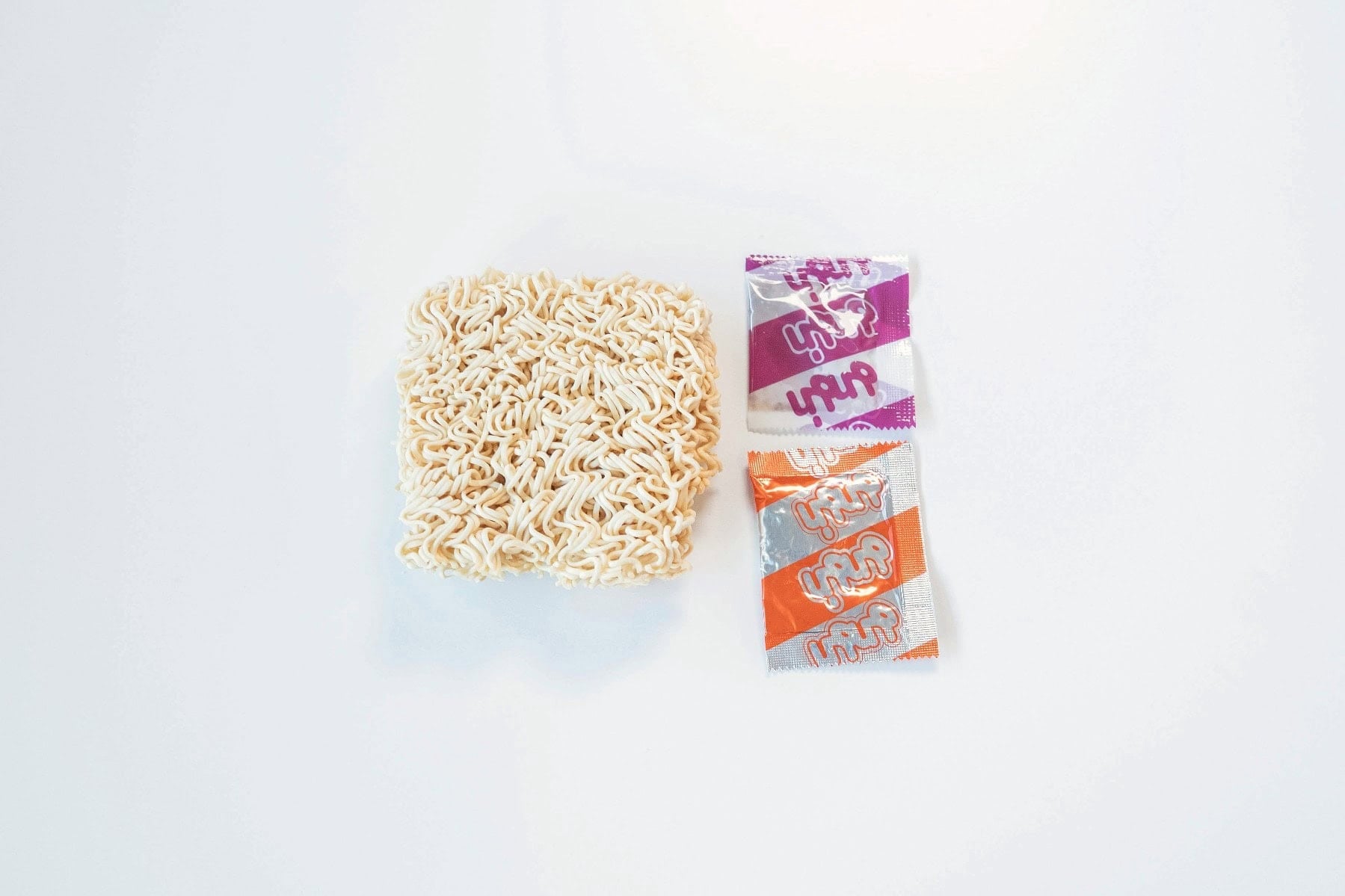 The contents of the MAMA Stew Beef Flavor Instant Noodles pack laid out on a white surface. A square block of thin wavy instant wheat noodles sits beside two seasoning sachets, one purple and white and one orange and white, both printed with Thai script.