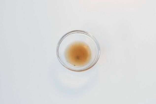 A small glass ramekin of the MAMA Stew Beef Flavor Instant Noodles broth on a white surface, showing a clear amber brown liquid with a few flecks of seasoning at the bottom.