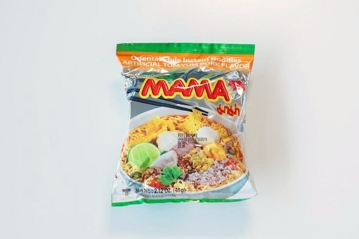 The front of the MAMA Tom Yum Pork Instant Noodles package on a white surface, showing the green and orange foil packaging with the red MAMA logo, Oriental Style Instant Noodles text, and a serving-suggestion photo of noodles with fish balls, lime, ground pork, chili, and cilantro in a red broth. Net Wt. 2.12 oz (60g).