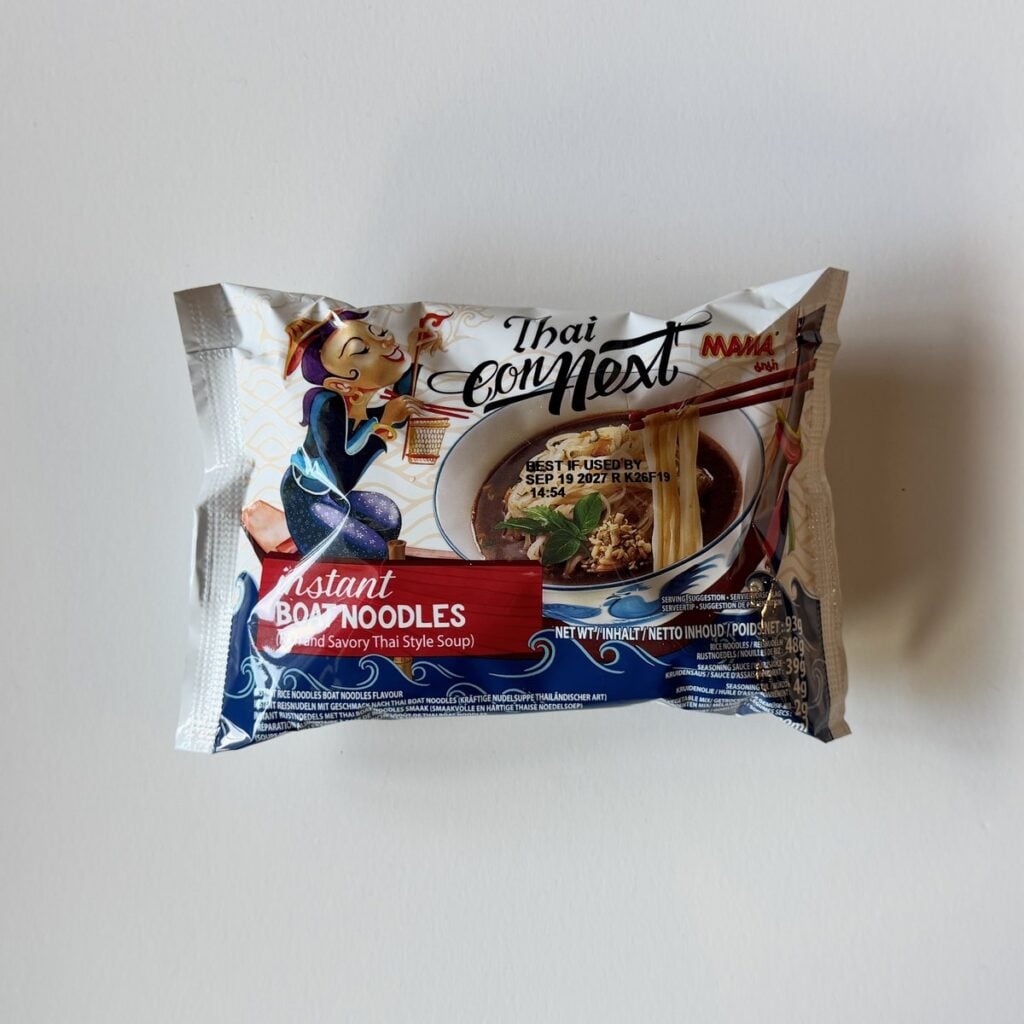 The MAMA Thai ConNext Instant Boat Noodles pack on a white table, with an illustrated woman holding chopsticks beside a bowl of dark broth and rice noodles under a red Instant Boat Noodles banner.