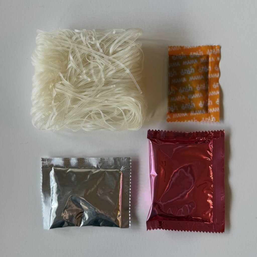 The contents of the MAMA Thai ConNext Instant Boat Noodles pack on a white surface: a block of flat rice noodles, a small orange seasoning packet, a silver dried garlic and herb packet, and a red seasoning sauce packet.