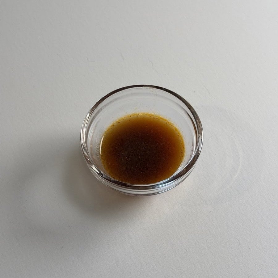 A small glass ramekin of the MAMA Thai ConNext Instant Boat Noodles broth on a white table, dark reddish brown with an orange ring of chili oil at the edge.