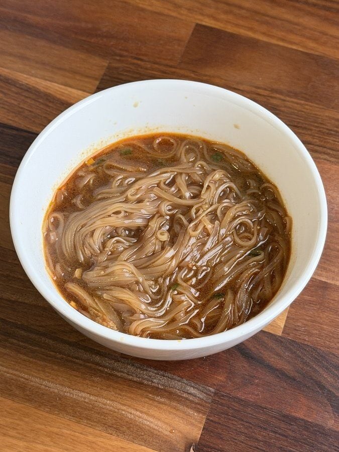A white bowl of MAMA Thai ConNext Instant Boat Noodles on a dark wood counter, flat rice noodles in a reddish brown broth with bits of green onion.