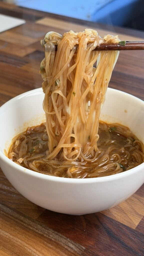 A close-up of flat rice noodles from the MAMA Thai ConNext Instant Boat Noodles lifted out of a white bowl with wooden chopsticks, coated in reddish brown broth.
