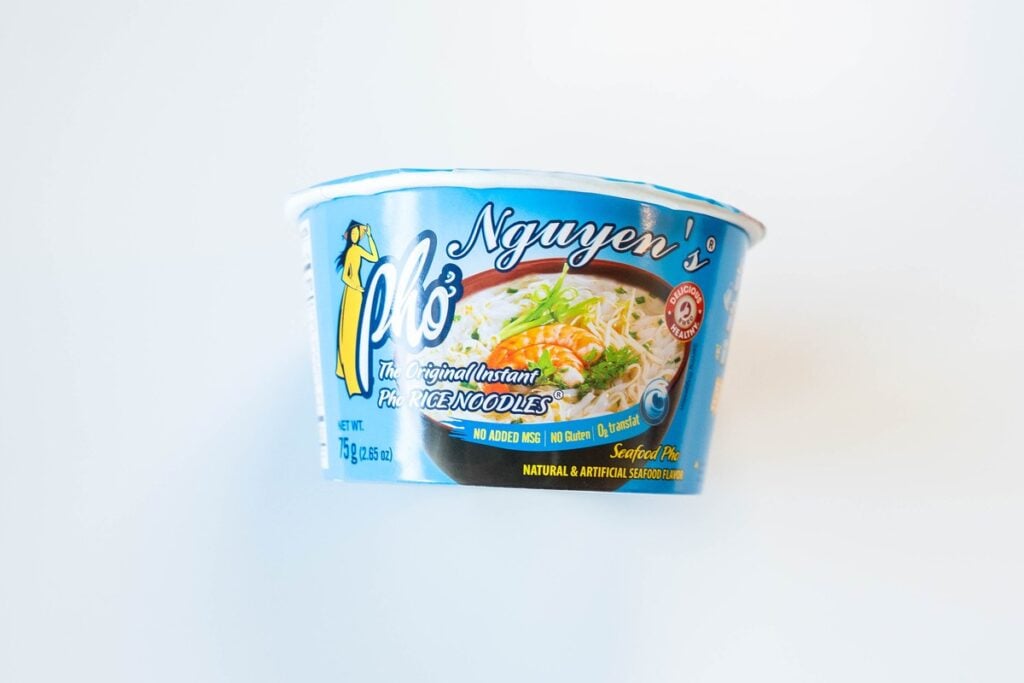The side of the Nguyen's Pho Instant Rice Noodles Seafood Flavor cup on a white surface, showing the Nguyen's Pho logo, an áo dài woman illustration, a bowl of pho with shrimp and herbs, and No Added MSG, No Gluten and 0g trans fat callouts.