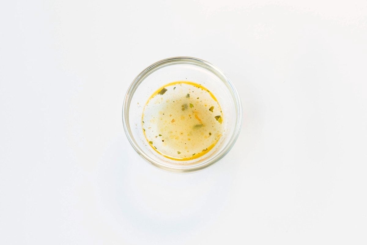 A small glass ramekin of the Nguyen's Pho Instant Rice Noodles Seafood Flavor broth on a white surface, showing a pale amber-yellow liquid with visible chili flecks and small pieces of dried green herbs.