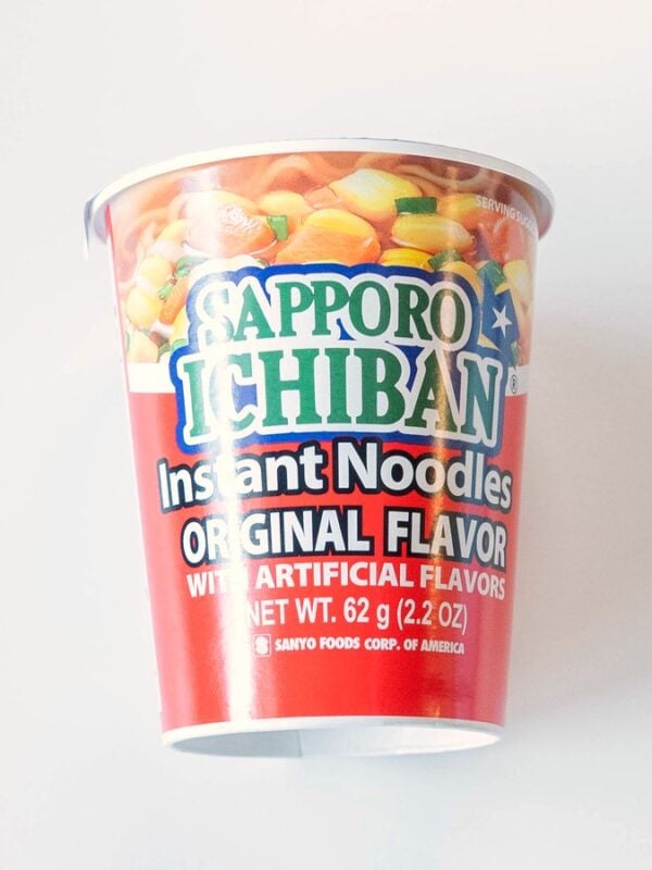 The red Sapporo Ichiban Original Flavor Cup on a white table, with the green and white Sapporo Ichiban logo over a photo of noodles and vegetables.