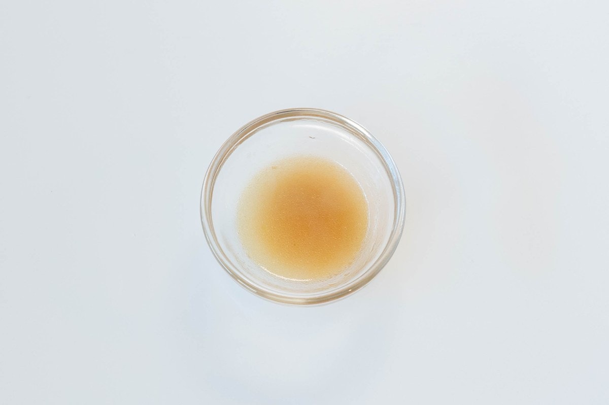 A small glass ramekin of the Ve Wong Dai Shi Ke Chicken Noodles with Rice Wine broth on a white surface, showing a clear light amber broth with a slight cloudy depth from the seasoning and rice wine.