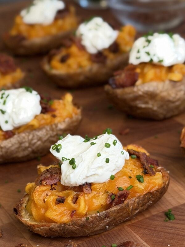Loaded ramen baked potato half on a butcher block, filled with orange sauced instant noodles and crumbled bacon, topped with a spoonful of sour cream and sliced chives, with more halves behind it.